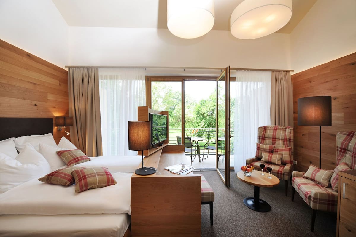 A hotel room with a bed, a TV, a seating area with a table, and a balcony with a view of greenery.