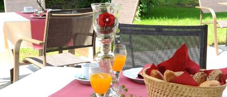 An outdoor dining table set with a red tablecloth, white plates, cutlery, a glass of orange juice, a basket of bread, and a vase with a red rose.