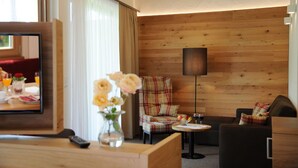 A hotel room with a wooden wall, a plaid armchair, a sofa, a small table with a wine glass, and a TV on a wooden stand. - Landgasthof Karolinenhöhe (Lichtenfels)