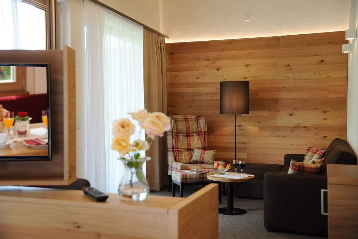 A hotel room with a wooden wall, a plaid armchair, a sofa, a small table with a wine glass, and a TV on a wooden stand.