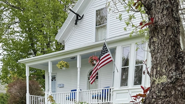 A white house with a porch, an American flag, and a tree with red leaves. - In-town 5 Bedroom Victorian With Heated Pool (Lubec)