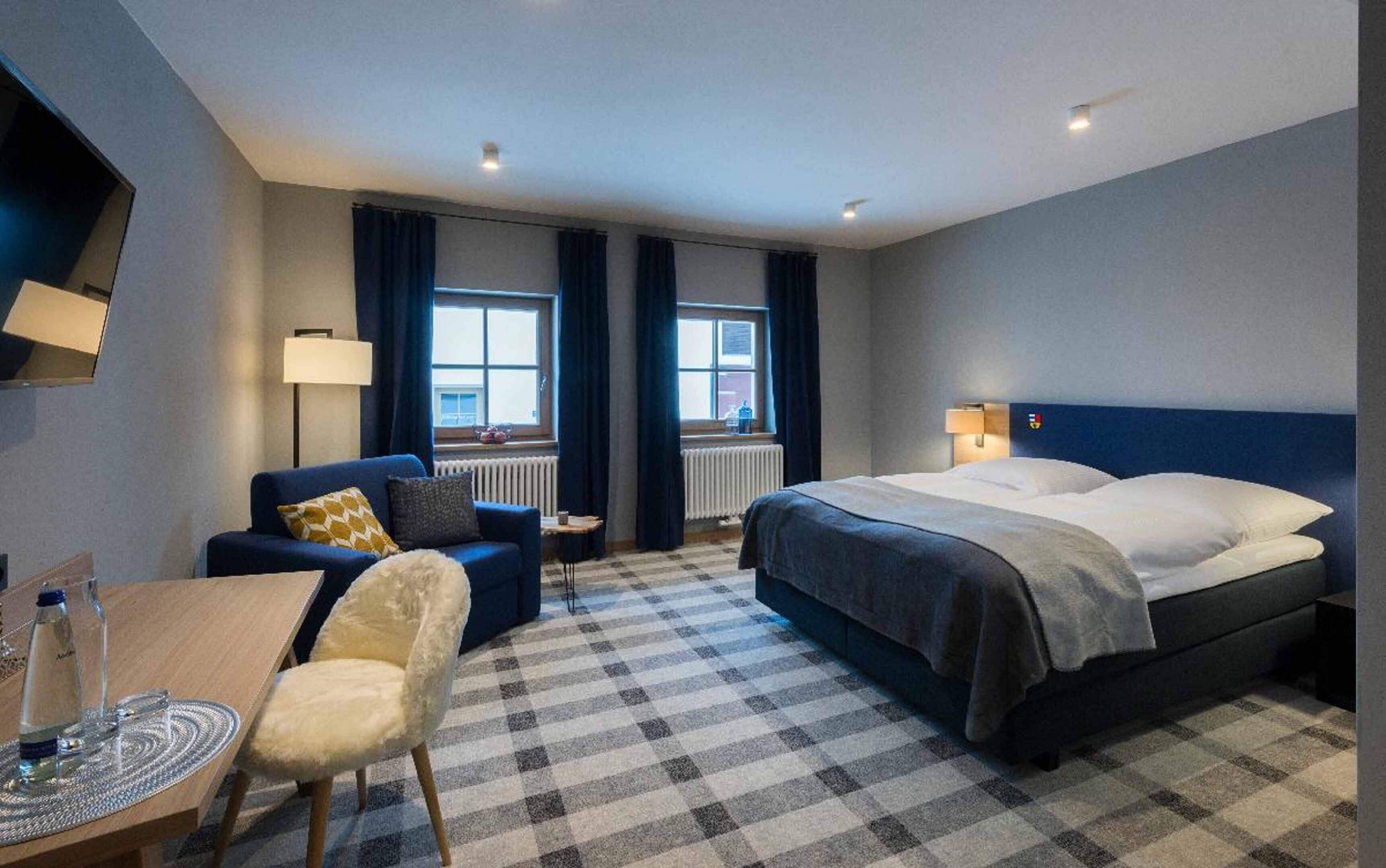 A hotel room with a checkered carpet, a bed with a blue headboard, a sofa with a yellow pillow, a small table with a water bottle, and two windows with curtains.