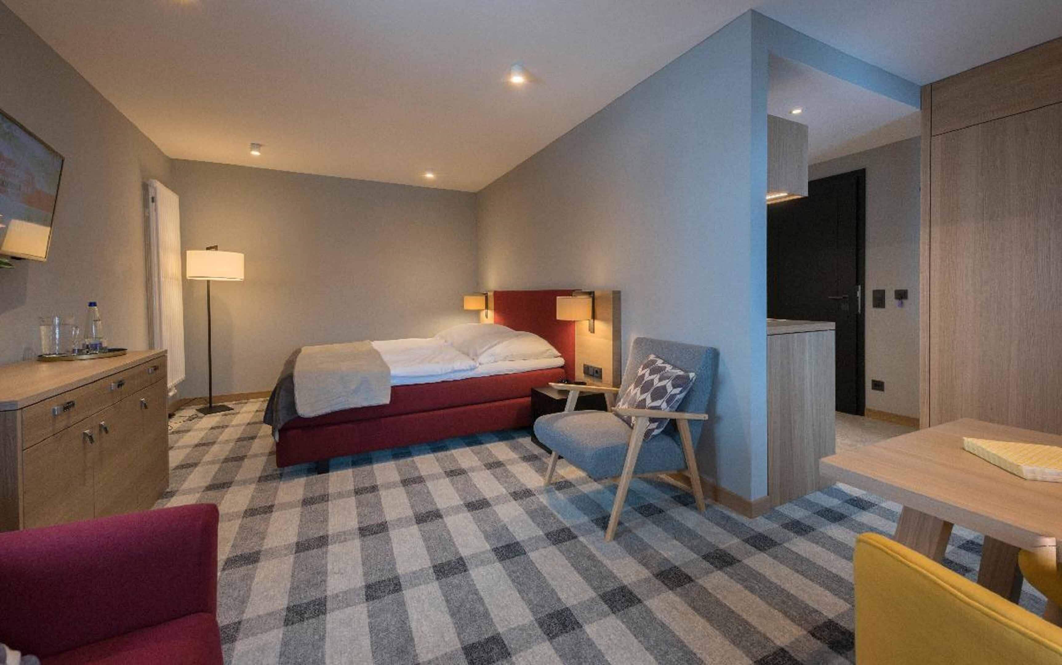 Une chambre d’hôtel avec un sol à carreaux, un lit avec une tête de lit rouge, un bureau avec une chaise, une armoire et une lampe.