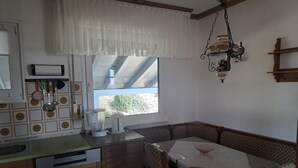A compact kitchen with wooden cabinets, a dining table with chairs, a window with a view, and a hanging light fixture. - Ferienwohnungen Alpenblick (Eppenschlag)