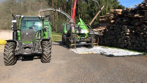 A green tractor with a front loader and a red crane in a rural setting with a pile of logs. - Schelterhof (Wunsiedel)