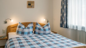 A bed with a checkered blanket, two pillows, a wooden headboard, two bedside lamps, a small framed picture on the wall, and a window with curtains. - Schelterhof (Wunsiedel)