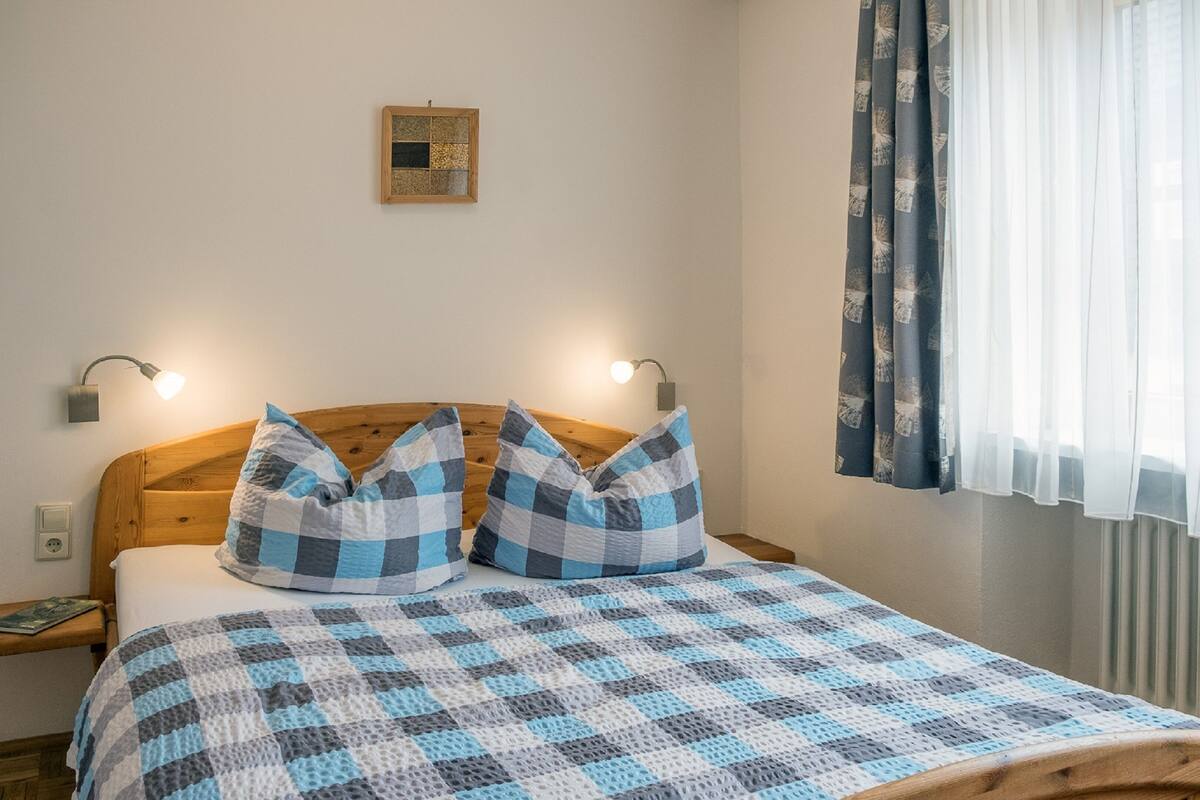 Ein Bett mit karierter Decke, zwei Kissen, einem hölzernen Kopfteil, zwei Nachttischlampen, einem kleinen gerahmten Bild an der Wand und einem Fenster mit Vorhängen.