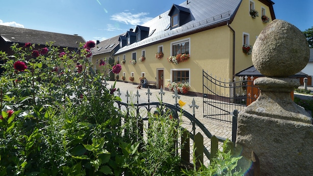 A yellow building with multiple windows and flower boxes, a stone bollard, and a paved walkway. - Schelterhof (Wunsiedel)