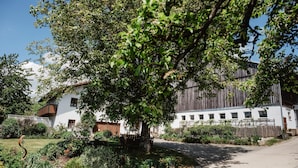 A white building with a balcony, surrounded by lush greenery and a tree. - Bio Ferienhof Winkler (Hutthurm)