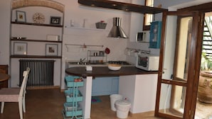 A compact kitchen with a built-in stove, microwave, and sink. There is a dining area with a chair and table, and a small trash bin. - Appartamento in Centro (Spoleto)