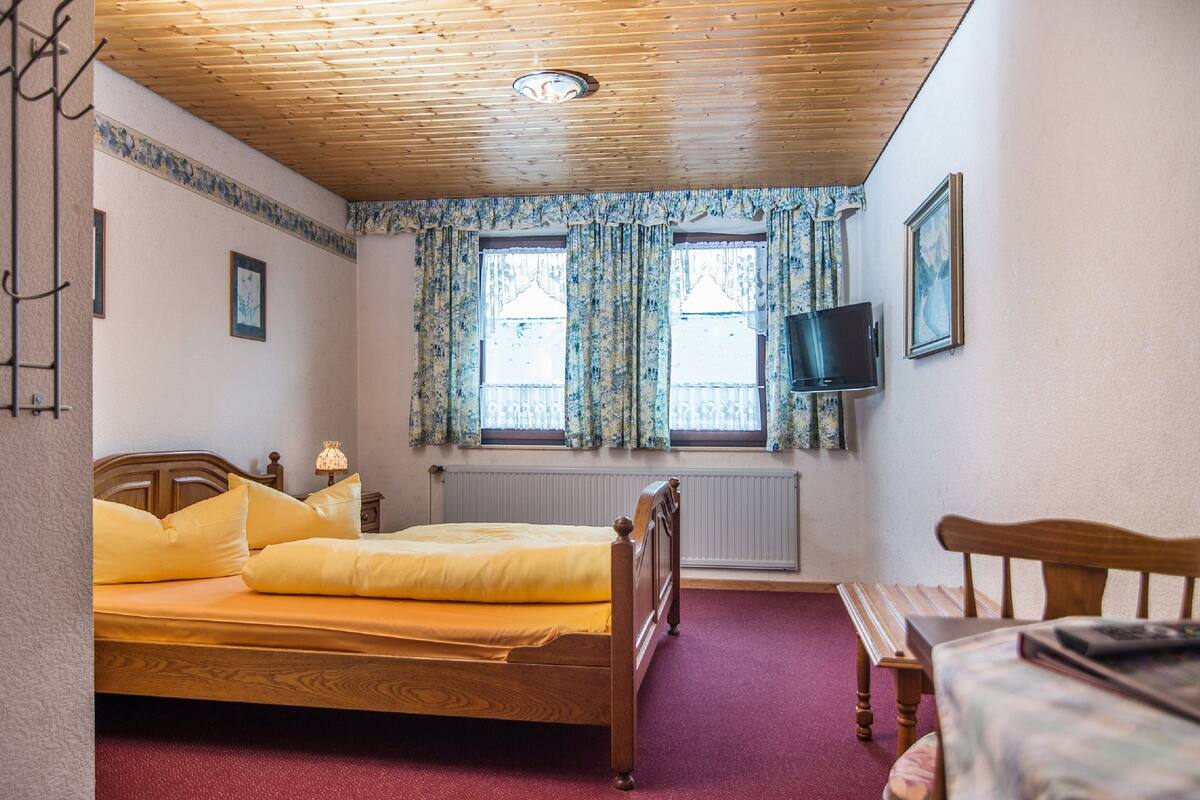 A bedroom with a wooden ceiling, a bed with yellow bedding, two windows with floral curtains, a bedside table, a TV mounted on the wall, and a radiator.