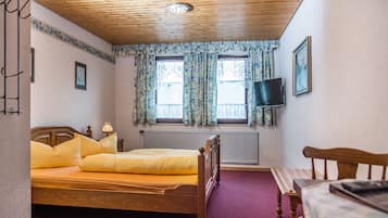 A bedroom with a wooden ceiling, a bed with yellow bedding, two windows with floral curtains, a bedside table, a TV mounted on the wall, and a radiator.