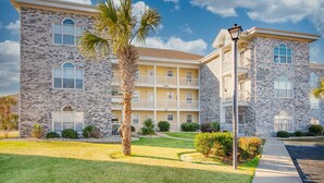 A multi-story building with a stone facade, balconies, and a well-maintained lawn. - 4695 Wild Iris Drive, #204 Magnolia Place 2 Bedroom Condo by RedAwning (Myrtle Beach)