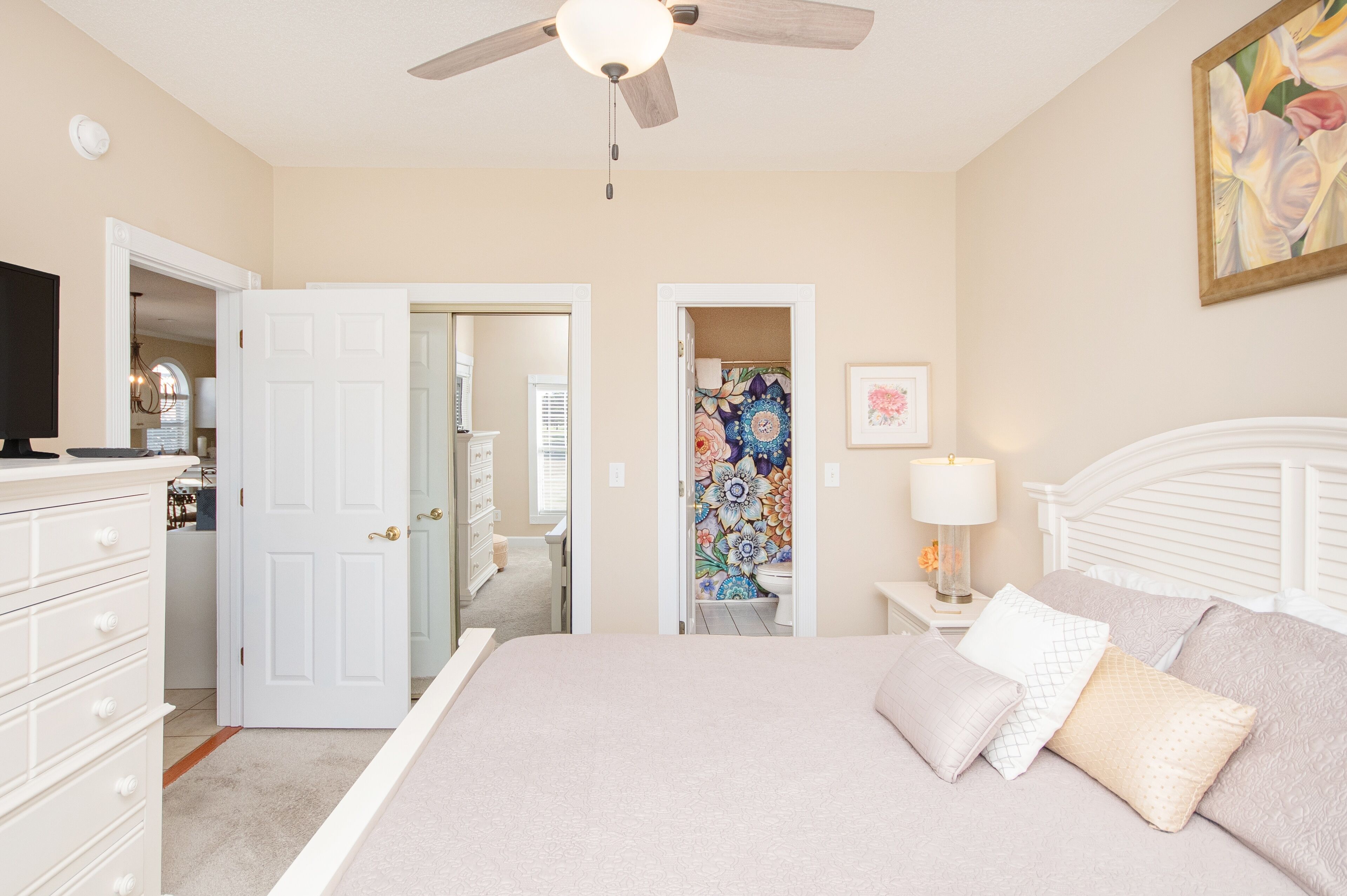 A bedroom with a bed, a dresser, a nightstand with a lamp, a ceiling fan, and a door leading to another room.