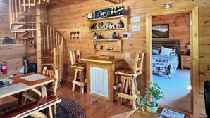 Interior de una cabaña con escalera de madera, comedor con mesa y sillas, pequeña zona de bar y un dormitorio acogedor visible a través de una puerta abierta.