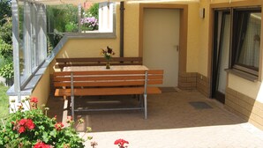 A covered outdoor seating area with a wooden bench and a small table, surrounded by potted plants and a door leading to an interior space. - Ferienwohnung Werner (Haundorf)