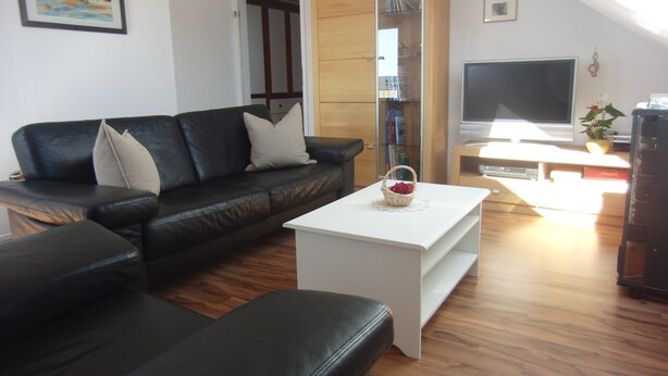 Una sala de estar con un sofá de cuero negro, una mesa de centro blanca con una cesta, un mueble de madera y un televisor.