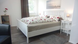 Un dormitorio con cama blanca, alfombra gris, mesita de noche con lámpara y ventana con cortinas.