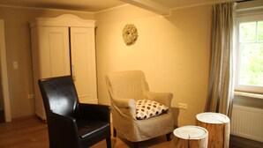A room with a wooden floor, a black chair, a beige armchair with a polka dot pillow, a wooden stool, and a white wardrobe. - Tauberlodge (Creglingen)