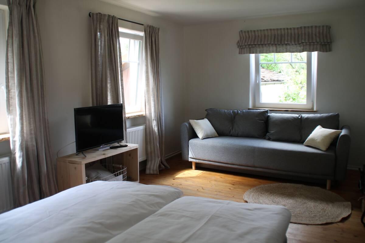 A bedroom with a bed, a TV on a wooden stand, a grey sofa, and a window with curtains.