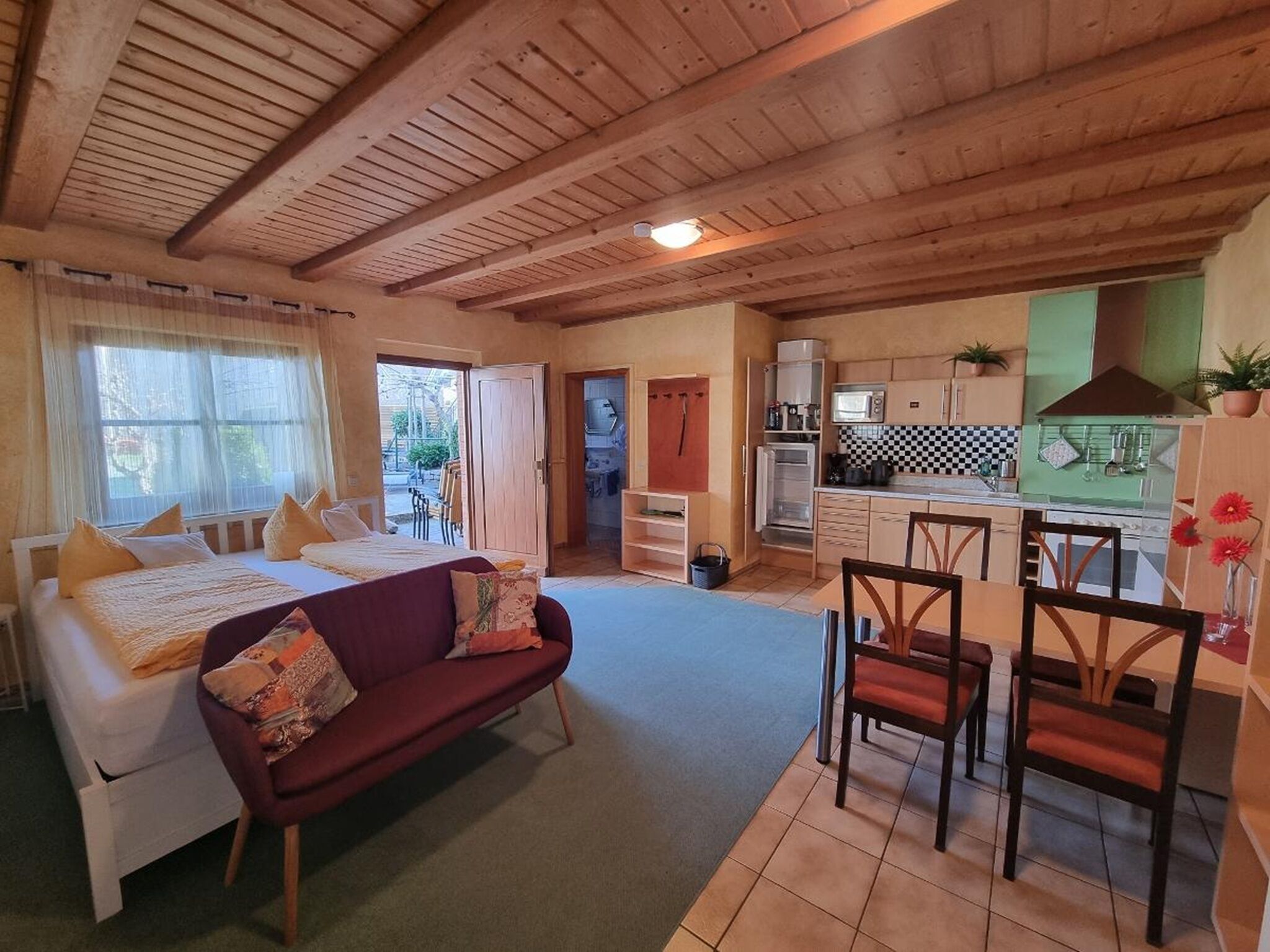 A cozy room with a bed, a sofa, a dining area with chairs, a kitchen with cabinets and appliances, and a view through an open door.