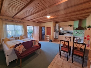 A cozy room with a bed, a sofa, a dining area with chairs, a kitchen with cabinets and appliances, and a view through an open door.