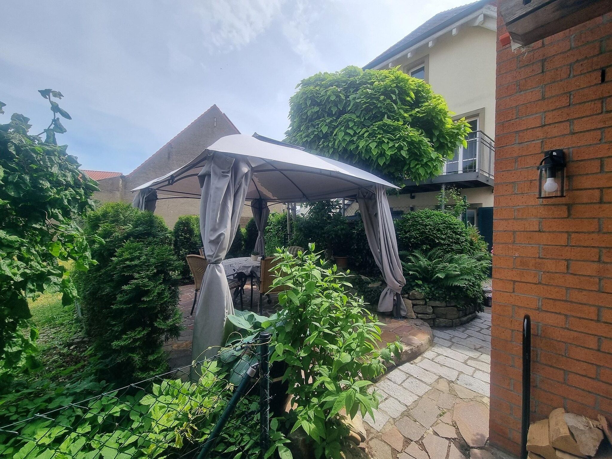A covered outdoor seating area with a table and chairs, surrounded by greenery and a brick wall.