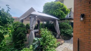A covered outdoor seating area with a table and chairs, surrounded by greenery and a brick wall.