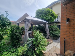 A covered outdoor seating area with a table and chairs, surrounded by greenery and a brick wall.