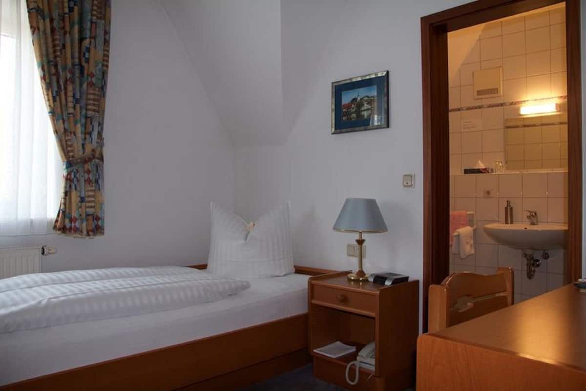 Une petite pièce propre, avec un lit simple, une table de chevet avec une lampe, une petite chaise et une salle de bain visible à travers une porte ouverte.