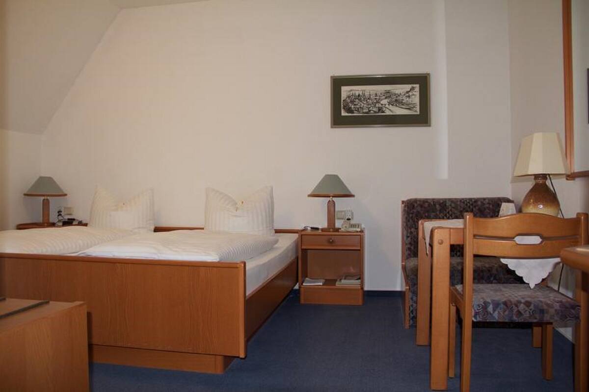 Une chambre d’hôtel avec un lit, deux tables de chevet avec des lampes, une chaise, une petite table et un tableau encadré au mur.