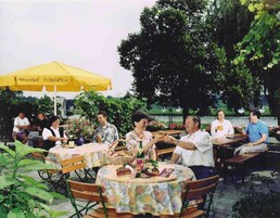Un área de comedor al aire libre con mesas y sillas, un paraguas amarillo y un cuerpo de agua al fondo.