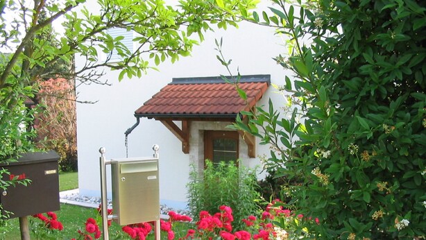 A white building with a red-tiled roof, surrounded by lush greenery and vibrant red flowers. - Ferienwohnung im Grünen (Illschwang)