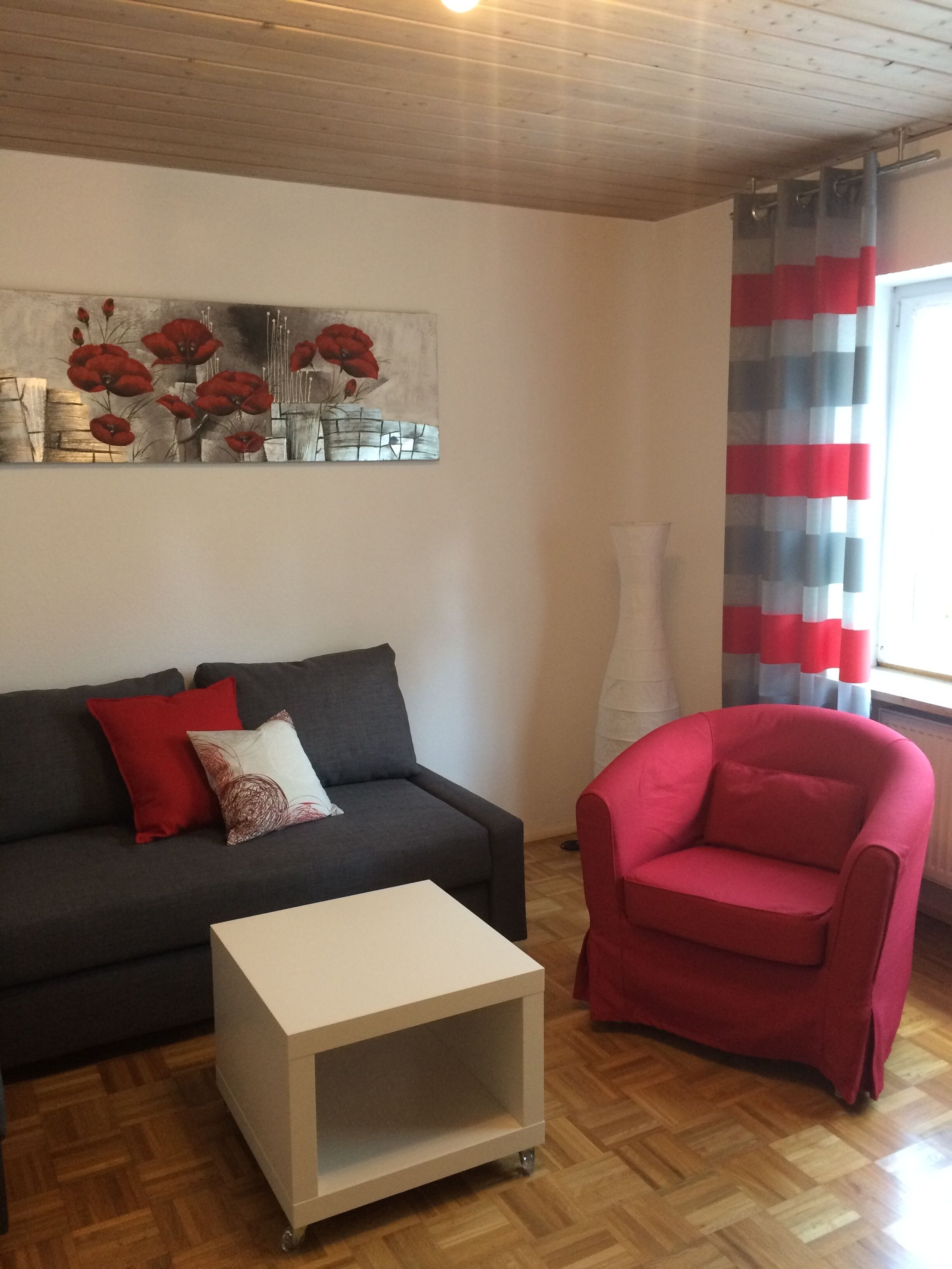 A living room with a dark grey sofa, a white coffee table, a red armchair, and a painting of red flowers on the wall.
