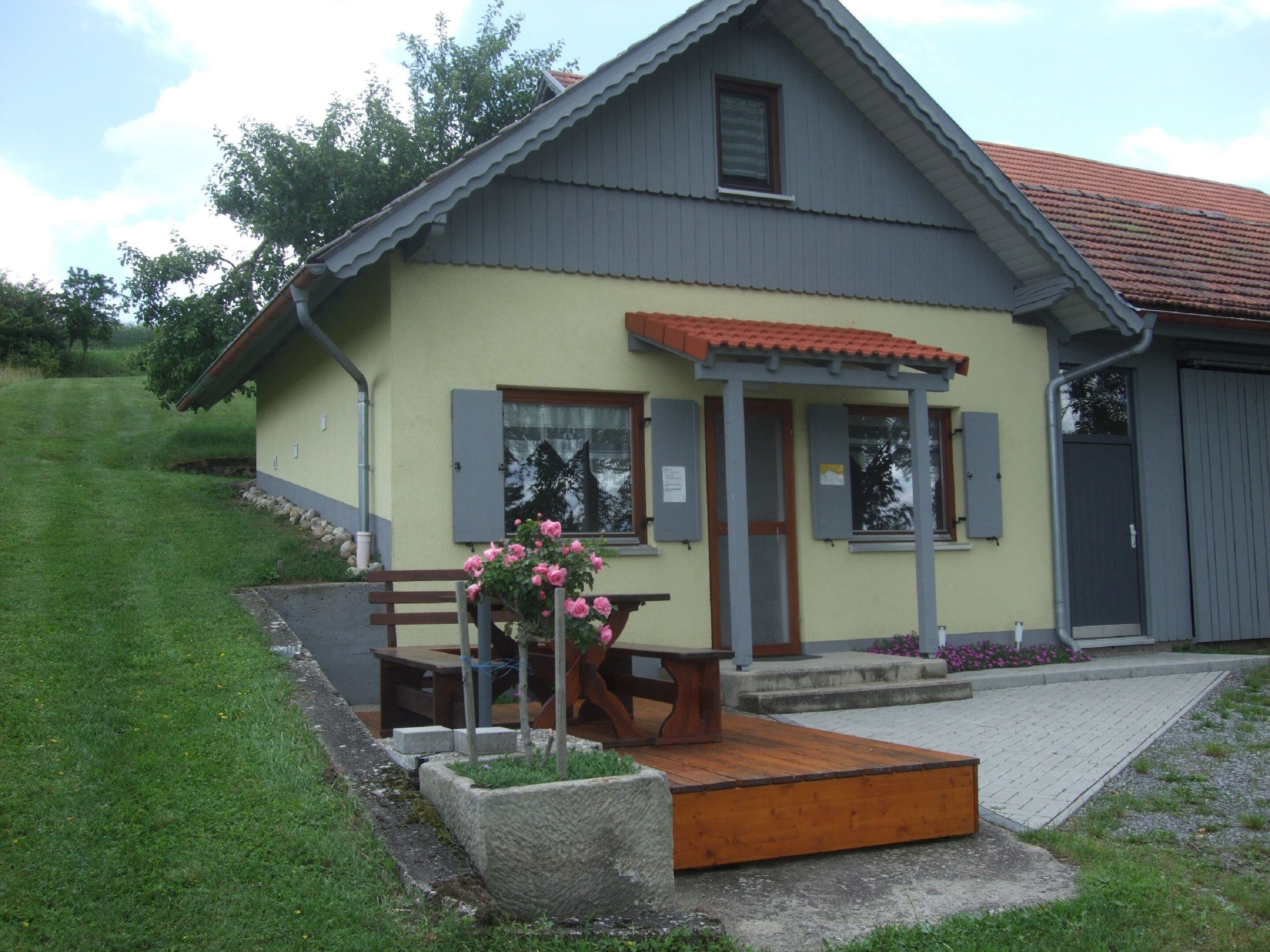 A two-story house with a sloped roof, a porch with a wooden bench, and a small garden with pink flowers.