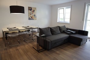 A modern living room with a grey sofa, a dining table with chairs, a hanging black lamp, and a window with blinds.
