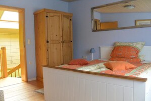A bedroom with a wooden wardrobe, a bed with orange bedding, a mirror on the wall, and a view of a wooden staircase.