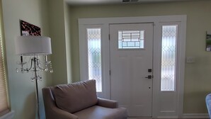 Een woonkamer met een beige bank, een glazen salontafel, een kroonluchter en een witte deur met geëtste glaspanelen.