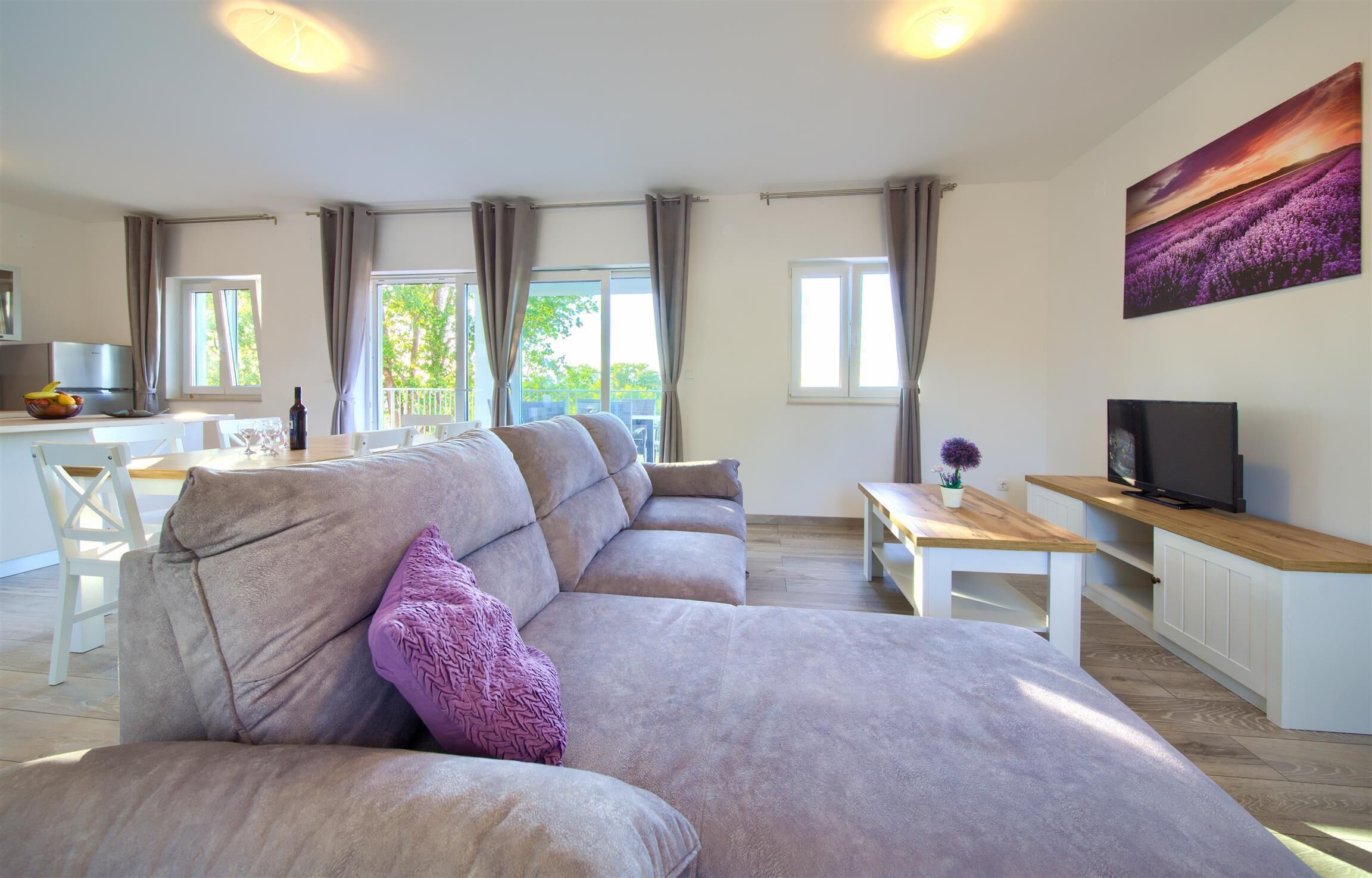 Una sala de estar moderna con un sofá beige grande, una mesa de centro de madera y un televisor sobre un mueble de madera. Hay dos ventanas con cortinas, un área de comedor con una mesa y sillas blancas, y un cuadro en la pared.