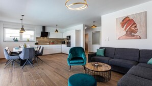 A modern living room with a sofa, teal armchair, and dining table. A kitchen is visible in the background with white cabinets and a black stove.