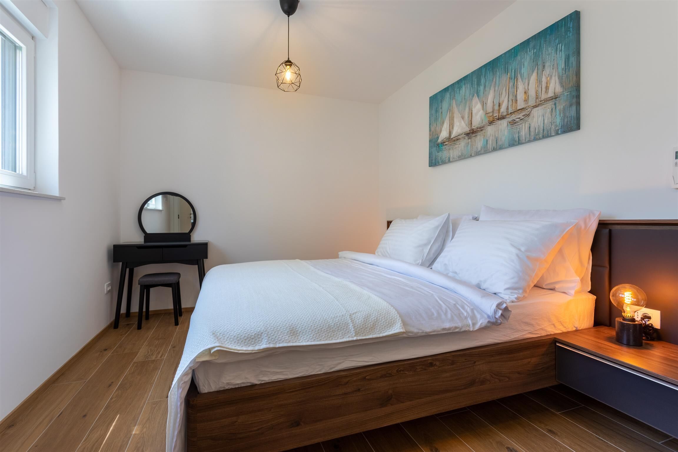 A bedroom with a wooden bed, a desk with a chair, a round mirror, a wall-mounted painting of sailboats, and a hanging light fixture.