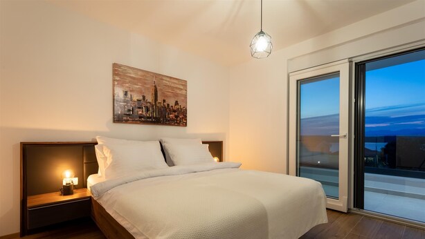 A modern bedroom with a cityscape painting, a bed with white linens, a nightstand with a lamp, and a large sliding glass door to the outside.