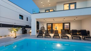 A modern two-story house with a swimming pool, outdoor seating, and a well-lit interior.