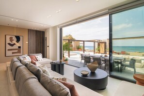 A modern living room with a large sofa, a coffee table, and a view of the sea through large windows. - Iris Beachfront Residence, Coastal City Living (Rethimno)