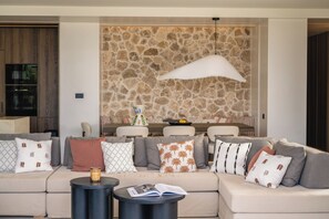 A modern living room with a stone wall, a sectional sofa, and a round coffee table. - Iris Beachfront Residence, Coastal City Living (Rethimno)