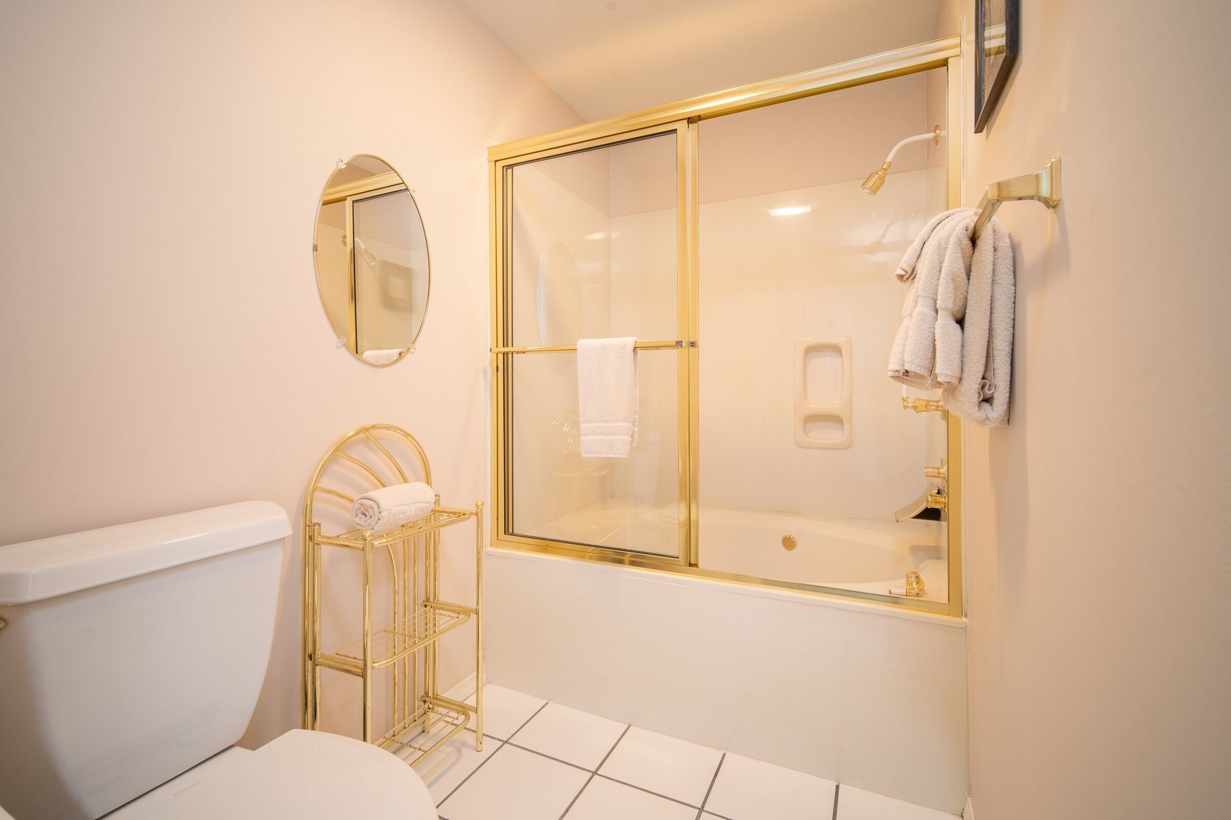 Et badeværelse med toilet, brusekabine i glas med guldframen, bøjle til håndklæder i guldfarve og et rundt spejl på væggen.