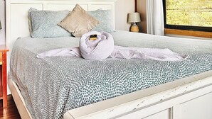 A bedroom with a bed, a headboard, a wall art of leaves, a window with curtains, and a bedside table.