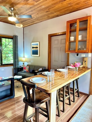 A dining area with a wooden table, chairs, a ceiling fan, a sofa, a lamp, and a window with curtains.