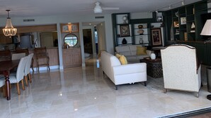 Amplia sala de estar con un sofá blanco, una mesa de comedor con sillas y una cocina con armarios de madera y una araña.