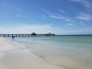 A beach with a pier extending into calm waters, a clear sky, and a few people walking along the shore.